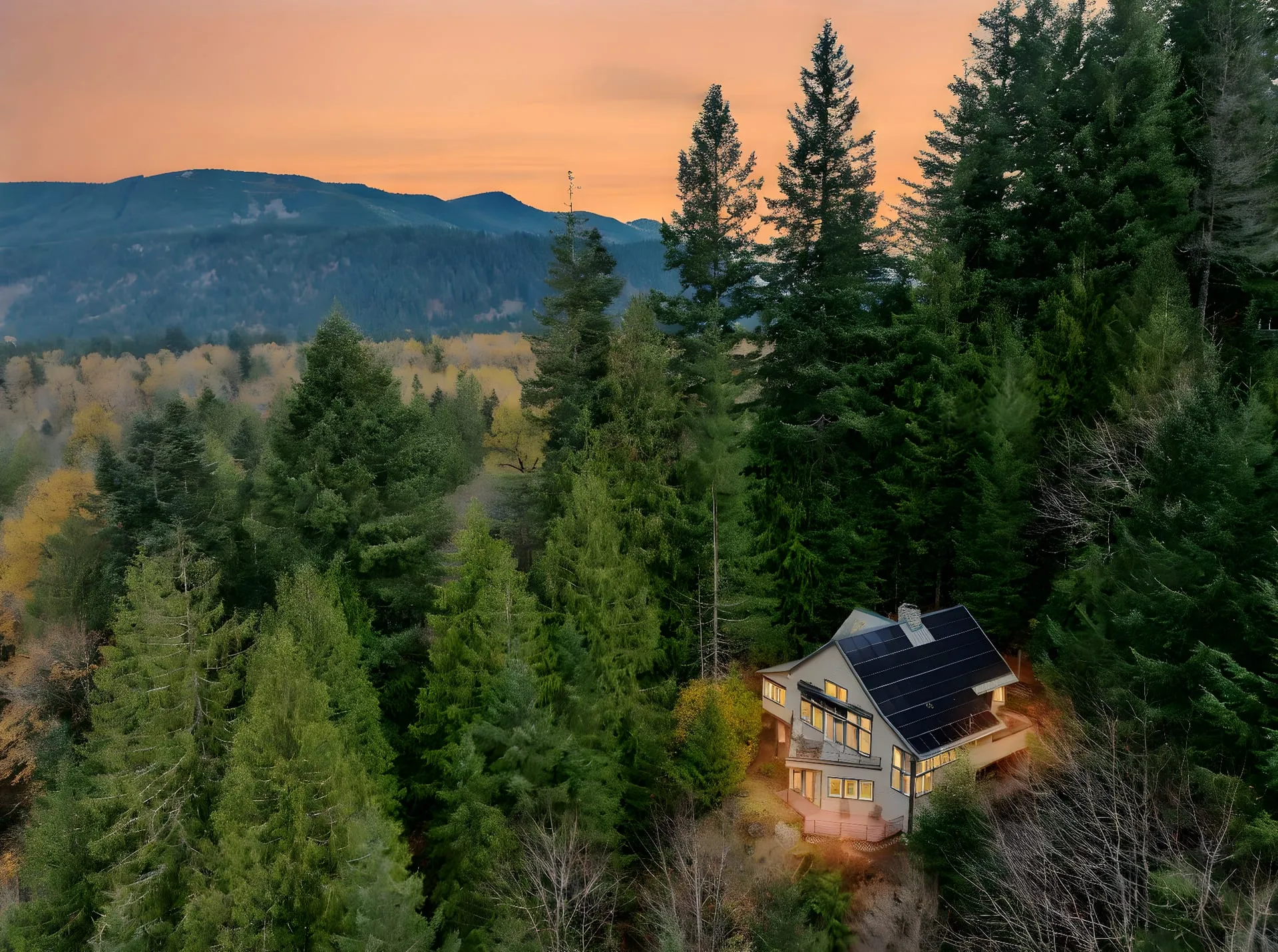 Aerial view of the home at dusk, tucked into tall evergreens with mountains beyond